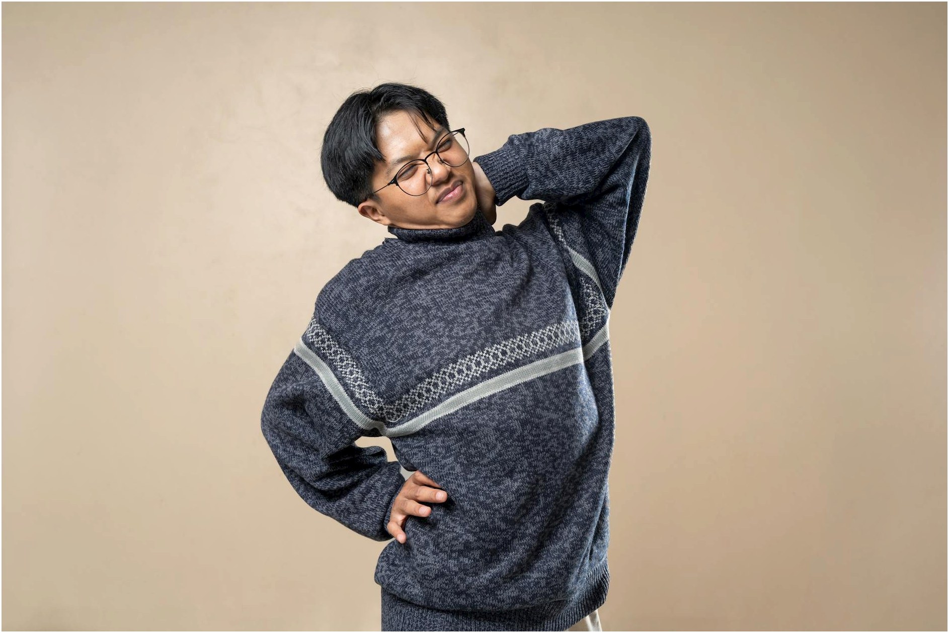 Asian man wearing eyeglasses and sweater stretching indoors, feeling body pain.