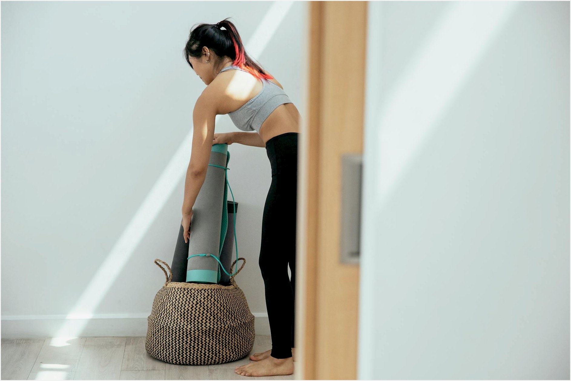 Side view full body of fit female in activewear standing near basket while holding mat in light room