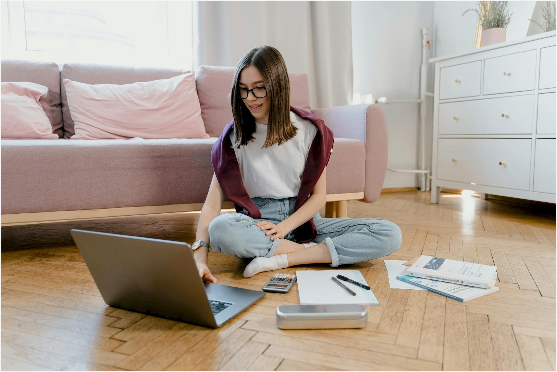 Teen girl studying online at home, sitting on the floor with a laptop and documents.