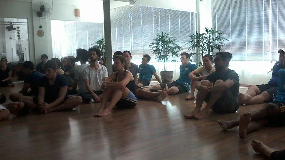 Alunos sentados em uma sala ampla de Pilates, com espaço livre no piso e equipamentos ao fundo