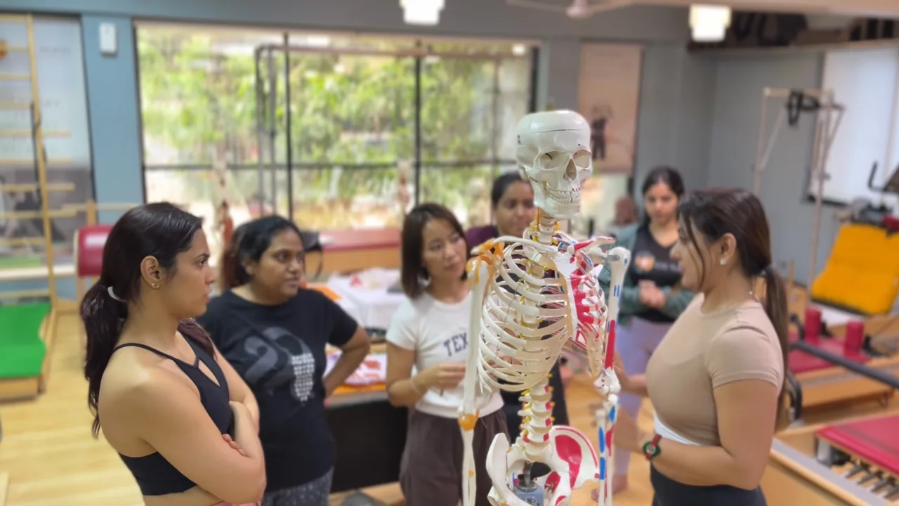 Instrutora orienta um grupo diante de um esqueleto anatômico em uma sala de Pilates com aparelhos ao fundo