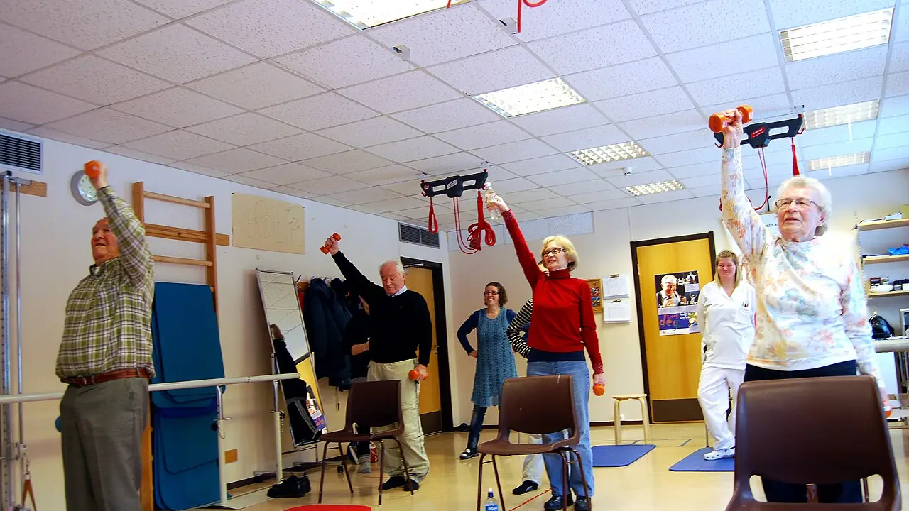 Pessoas idosas realizando exercício de força com halteres leves, com apoio de cadeiras.