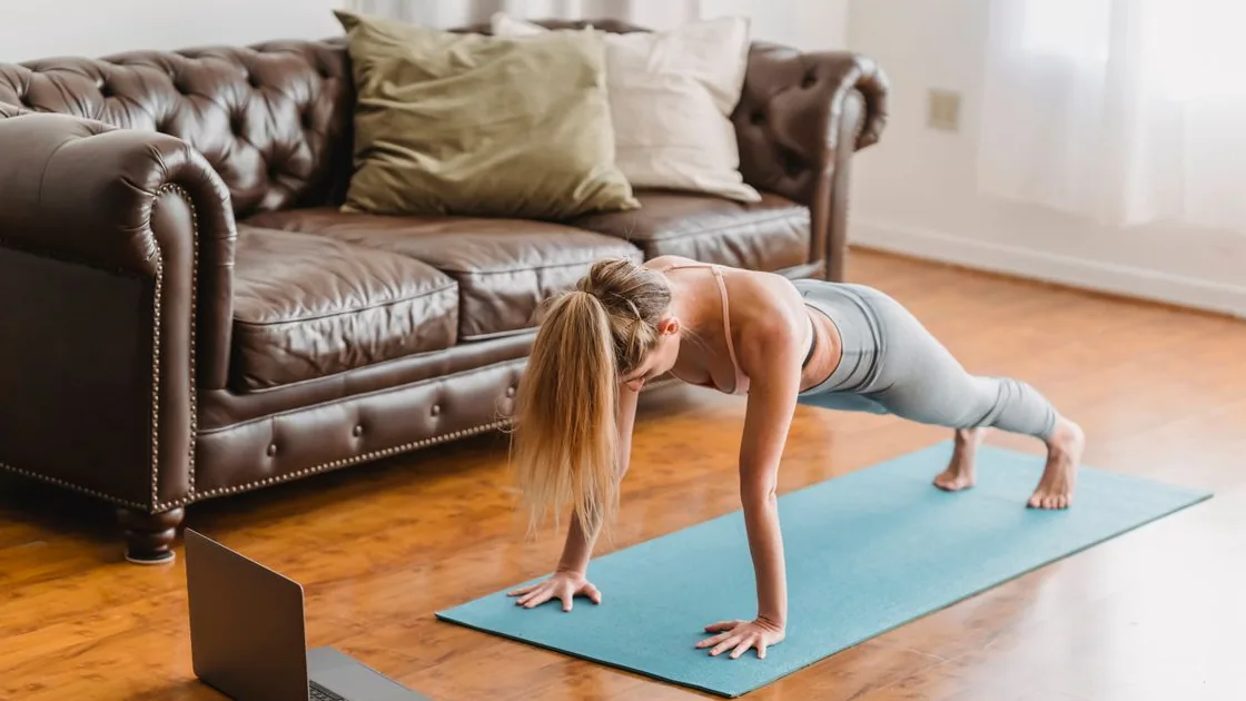 Pessoa realizando exercício no solo sobre um colchonete, com notebook ao lado, em ambiente doméstico