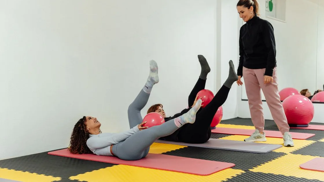 Aula de Pilates no solo com instrutora orientando alunas durante exercícios com bola