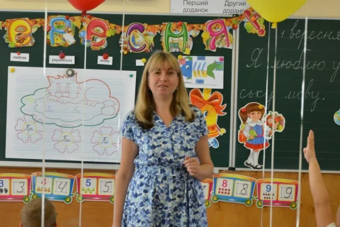 Professora em sala de aula diante do quadro, com alunos ao redor.