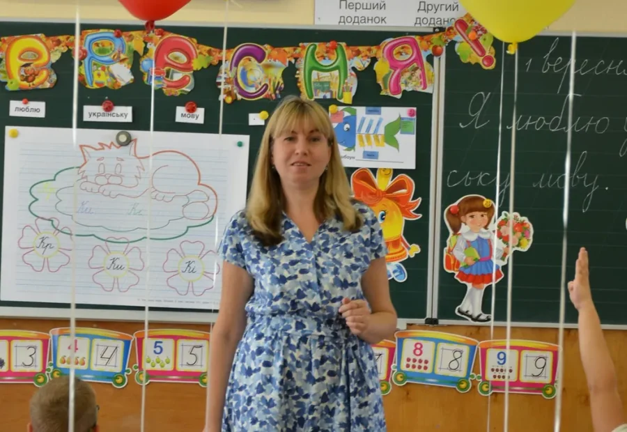 Professora em sala de aula diante do quadro, com alunos ao redor.
